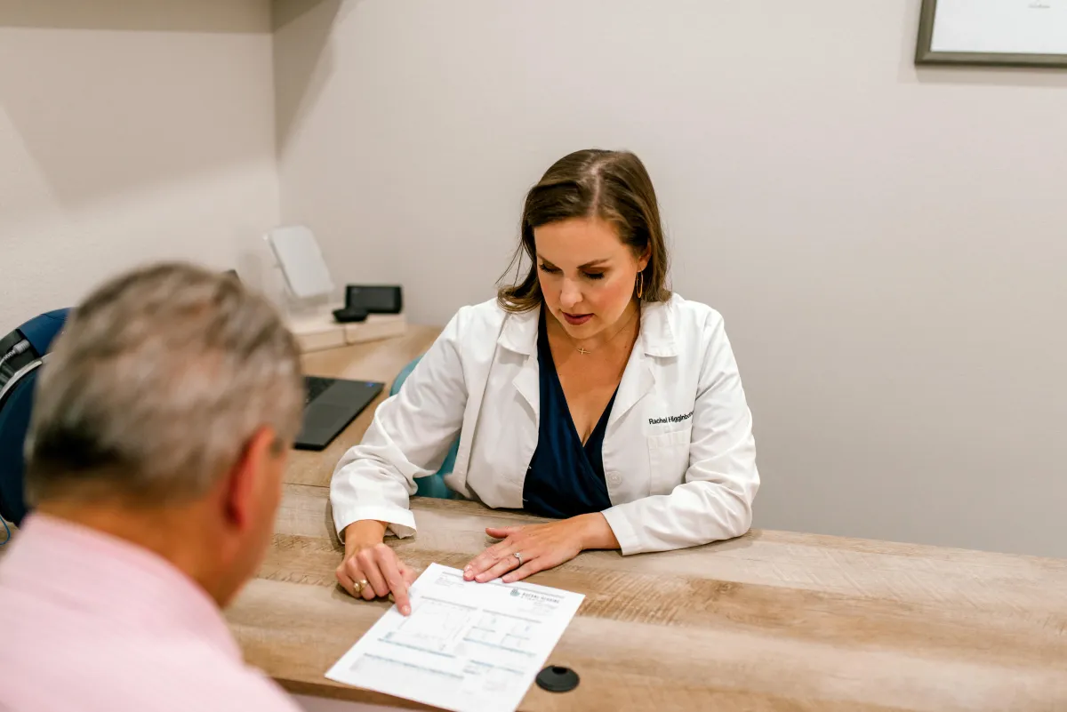 Audiologist pointing to a document while consulting an older male patient across a wooden desk.