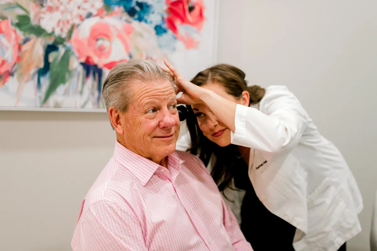 Audiologist in a white coat examining an older man's scalp in a clinic with floral artwork in the background.