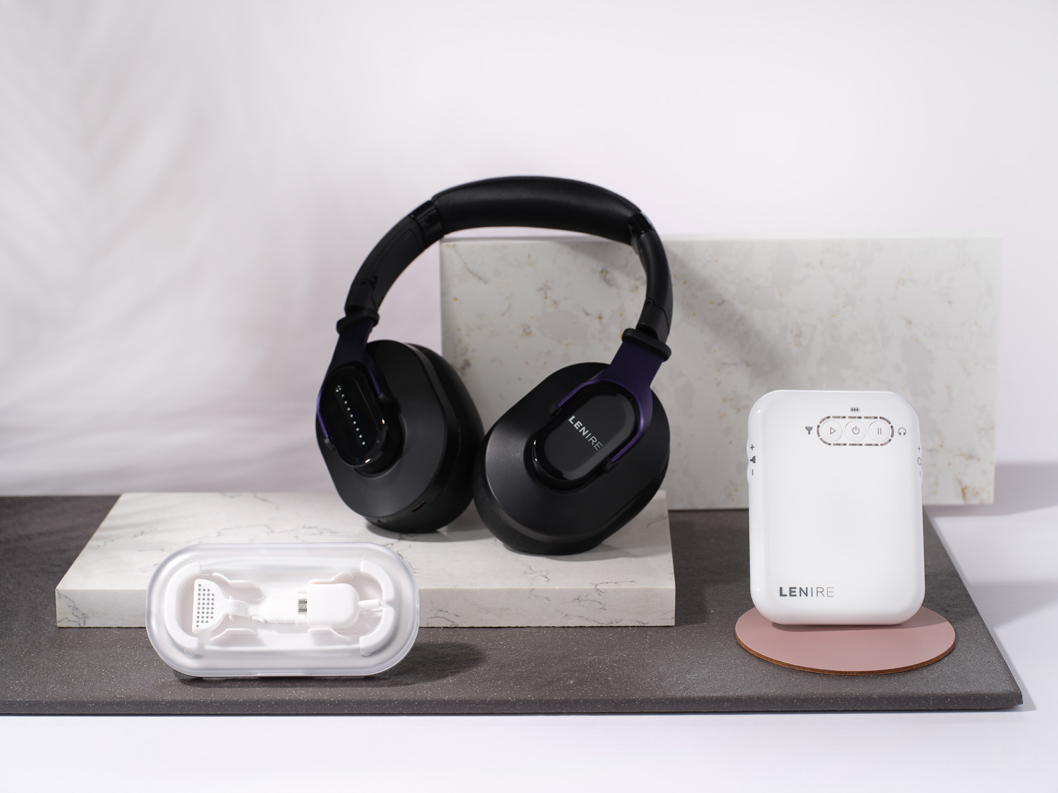 Lenire-branded black wireless over-ear headphones, a white control device, and a small white mouthpiece in a clear case displayed on marble and dark surfaces.