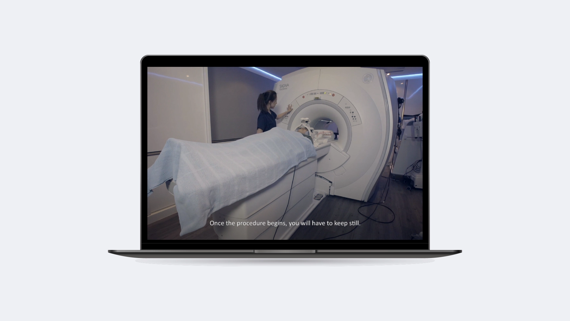 Patient lying under a blanket on an MRI scanner bed with a technician operating the machine, with the text 'Once the procedure begins, you will have to keep still.'