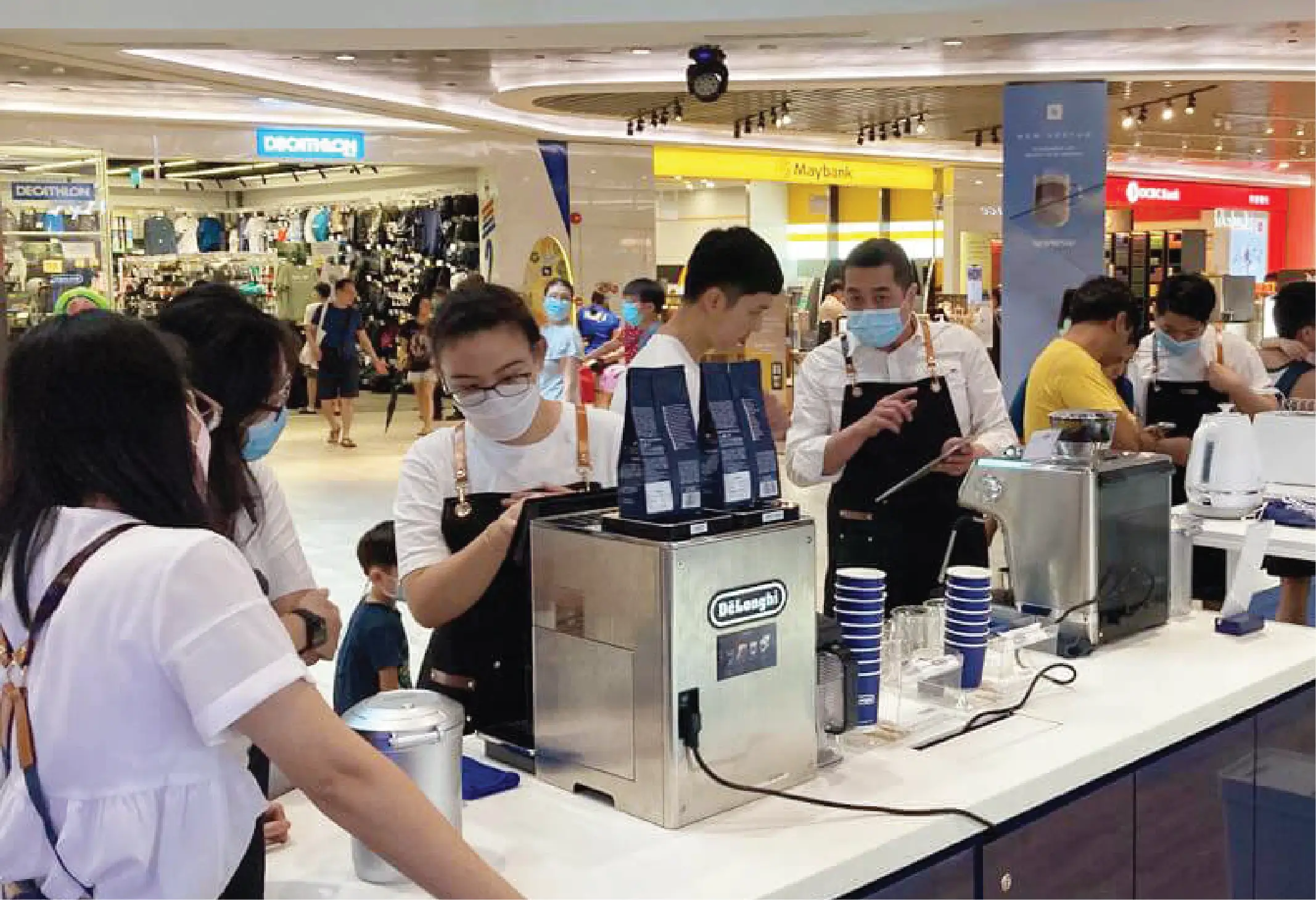 Crowd gathered around a coffee bar counter with DeLonghi appliances. Brand Ambassadors interact with customers, showcasing beverages in a lively retail environment.