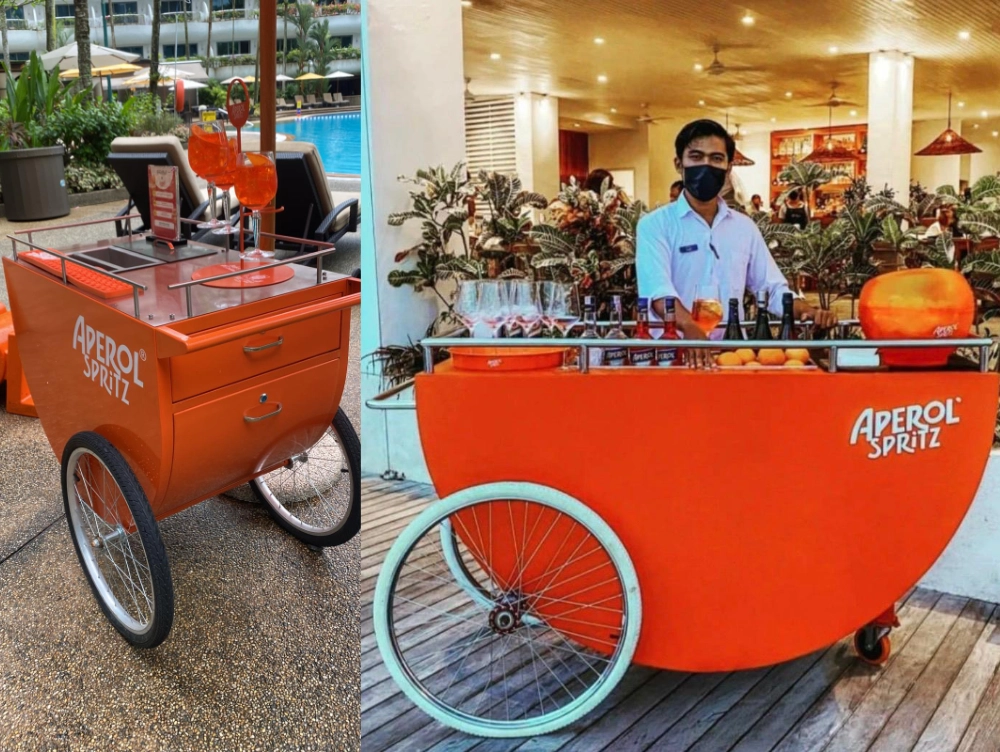 Orange Aperol Spritz mobile carts with glasses and bottles by a pool and a bartender indoors.