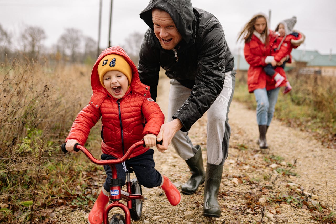 Une famille jouent dehors. Le papa aide son jeune fils à faire du vélo et la maman porte un enfant dans ses bras