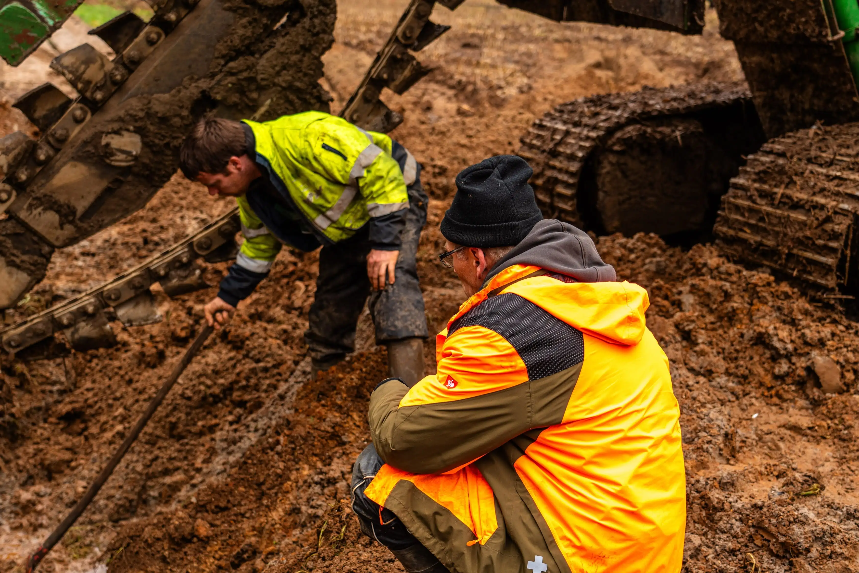 Two men in yellow jackets are kneeling in mud.