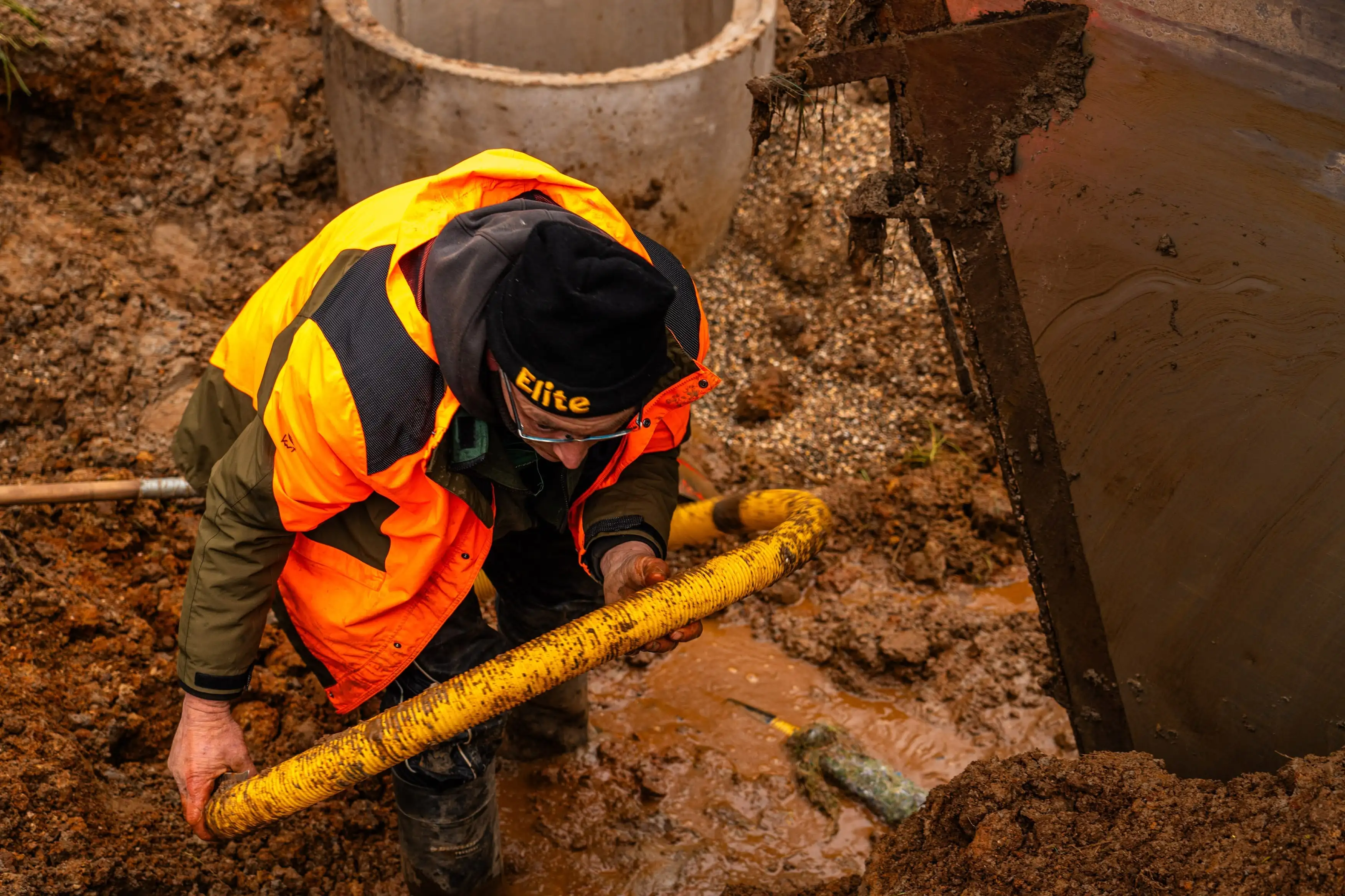 A man in a yellow jacket is bending over to pick up a yellow pipe.