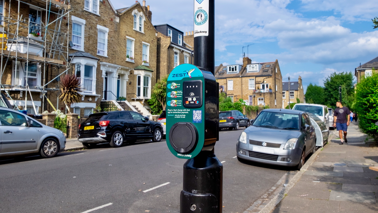 Zest on-street EV charging point mounted on a lamppost in a residential street in Hackney, London, with parked cars and Victorian-style houses in the background.