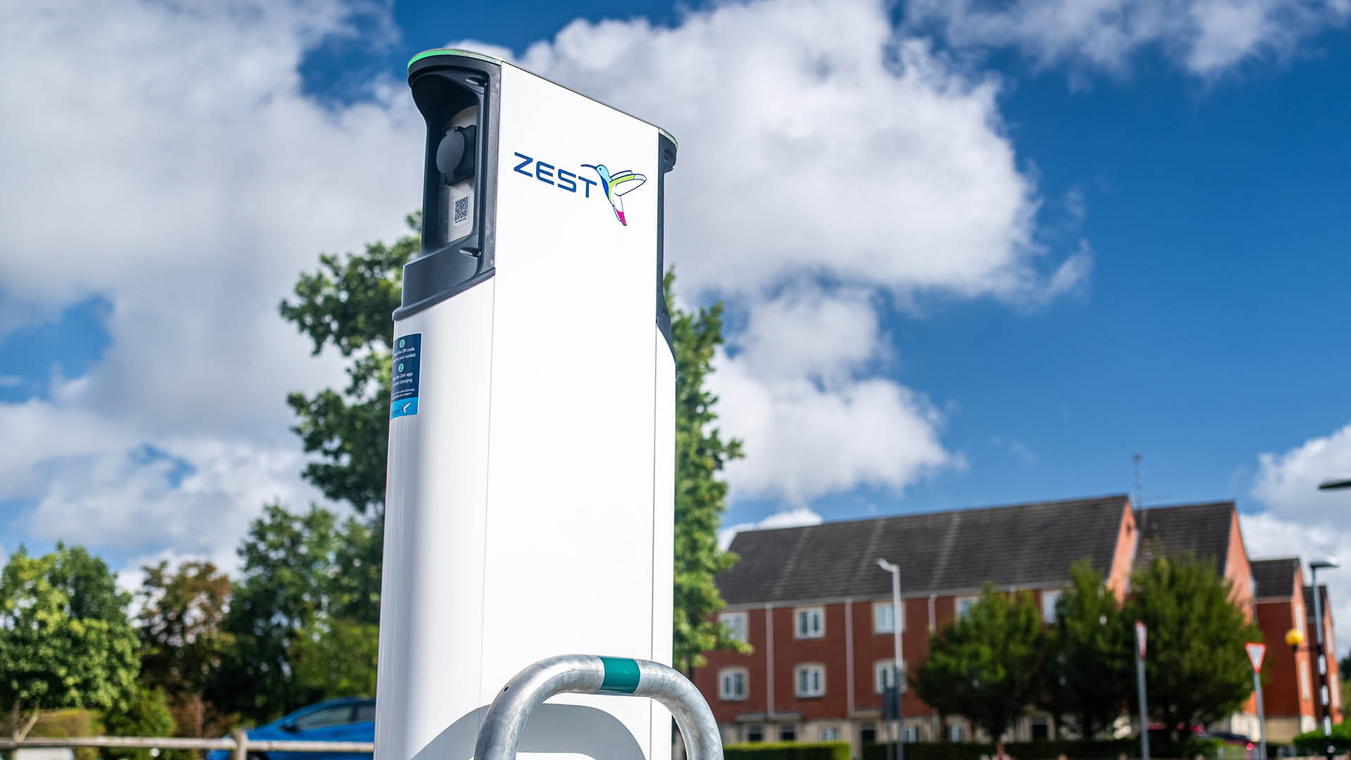 A Zest-branded electric vehicle charging point installed on a residential street, with houses, trees, and a blue sky with clouds in the background.