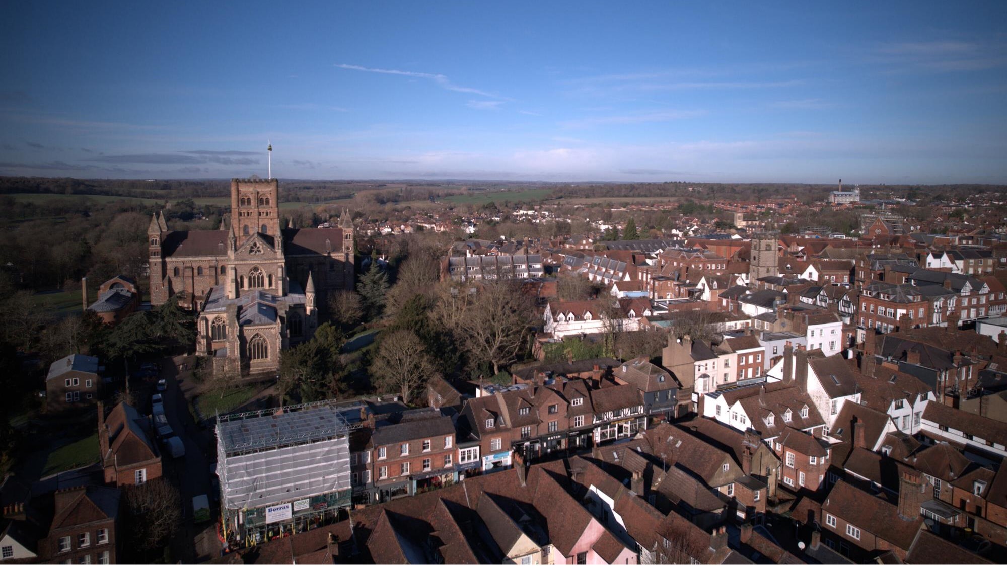 Aerial view of St Albans, Hertfordshire, showing the historic city centre and St Albans Cathedral, with surrounding homes, countryside and blue skies in the background.