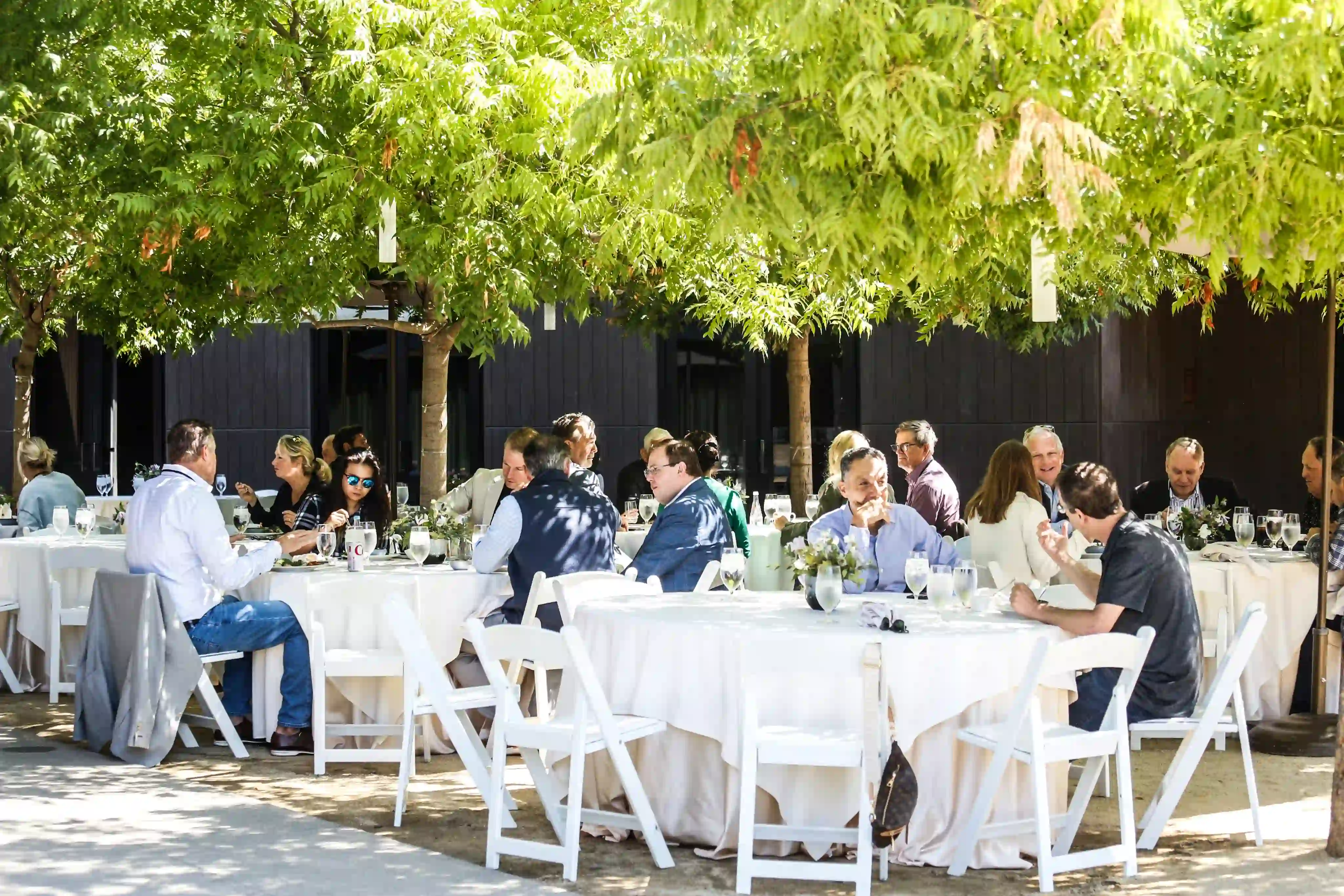 Image of a dinner at winery with green trees in the background, tents to cover the sun. Guests sitting in white chairs