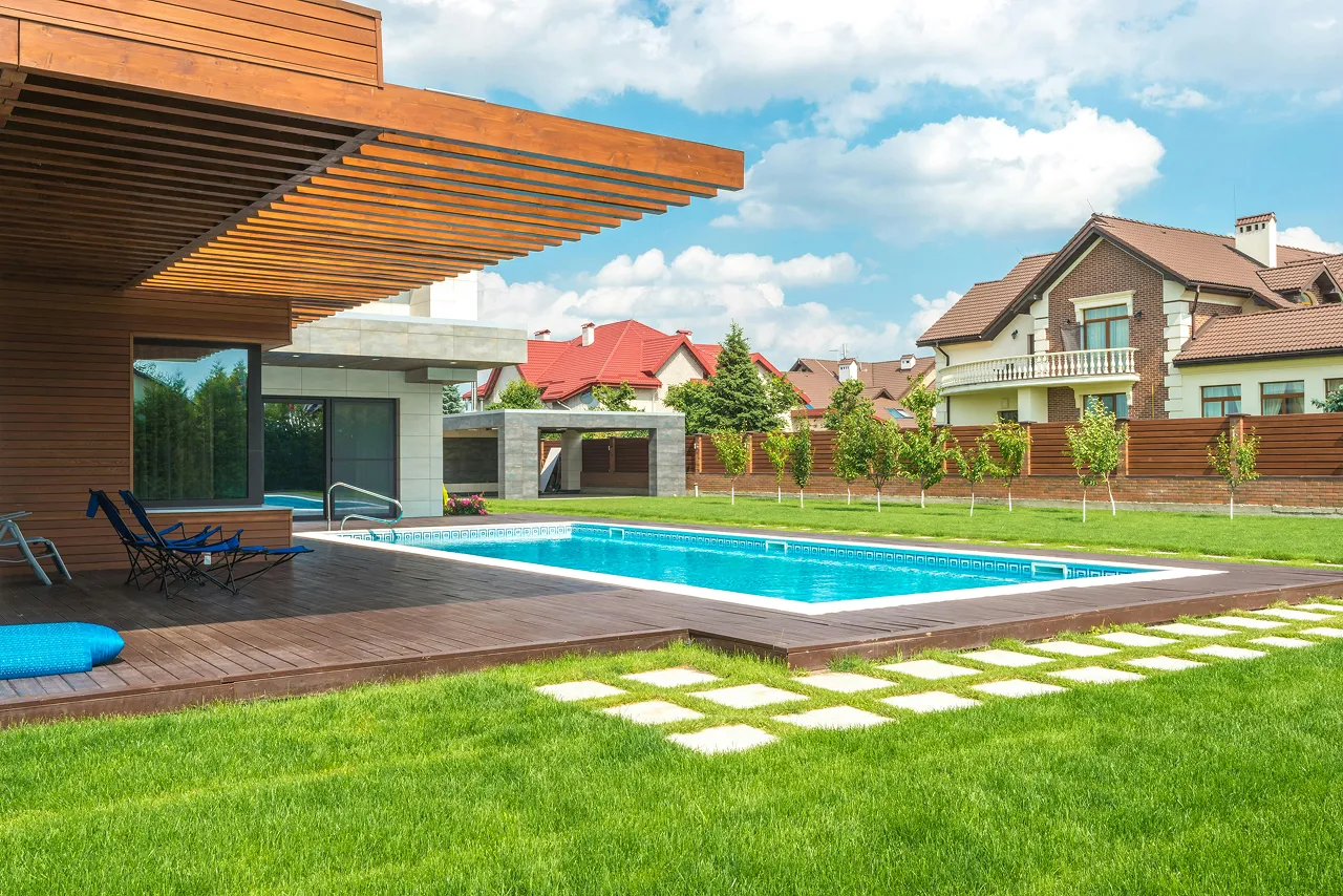 Modern house backyard with a blue swimming pool, wooden deck, green lawn, and clear sky.