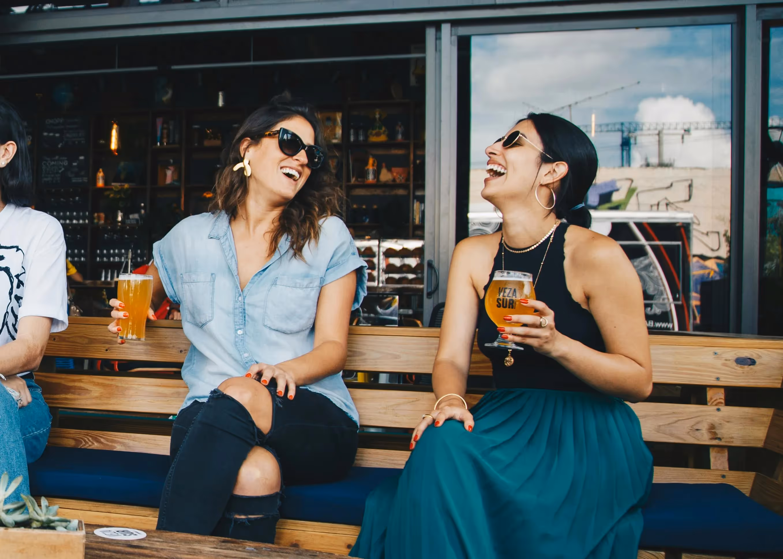 Two happy women laughing and enjoying drinks outdoors at a bar or restaurant, representing the vibrant social life and local attractions near aQui355 apartments in Salt Lake City, Utah.