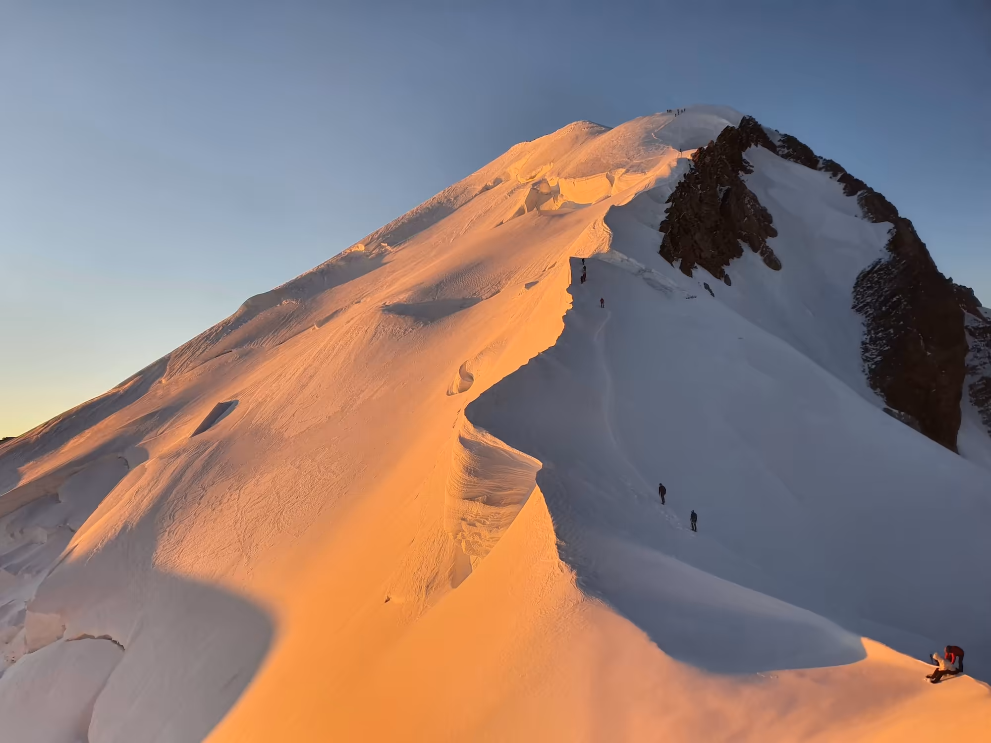 Sunlit snow-covered mountain peak with climbers ascending under a clear blue sky.