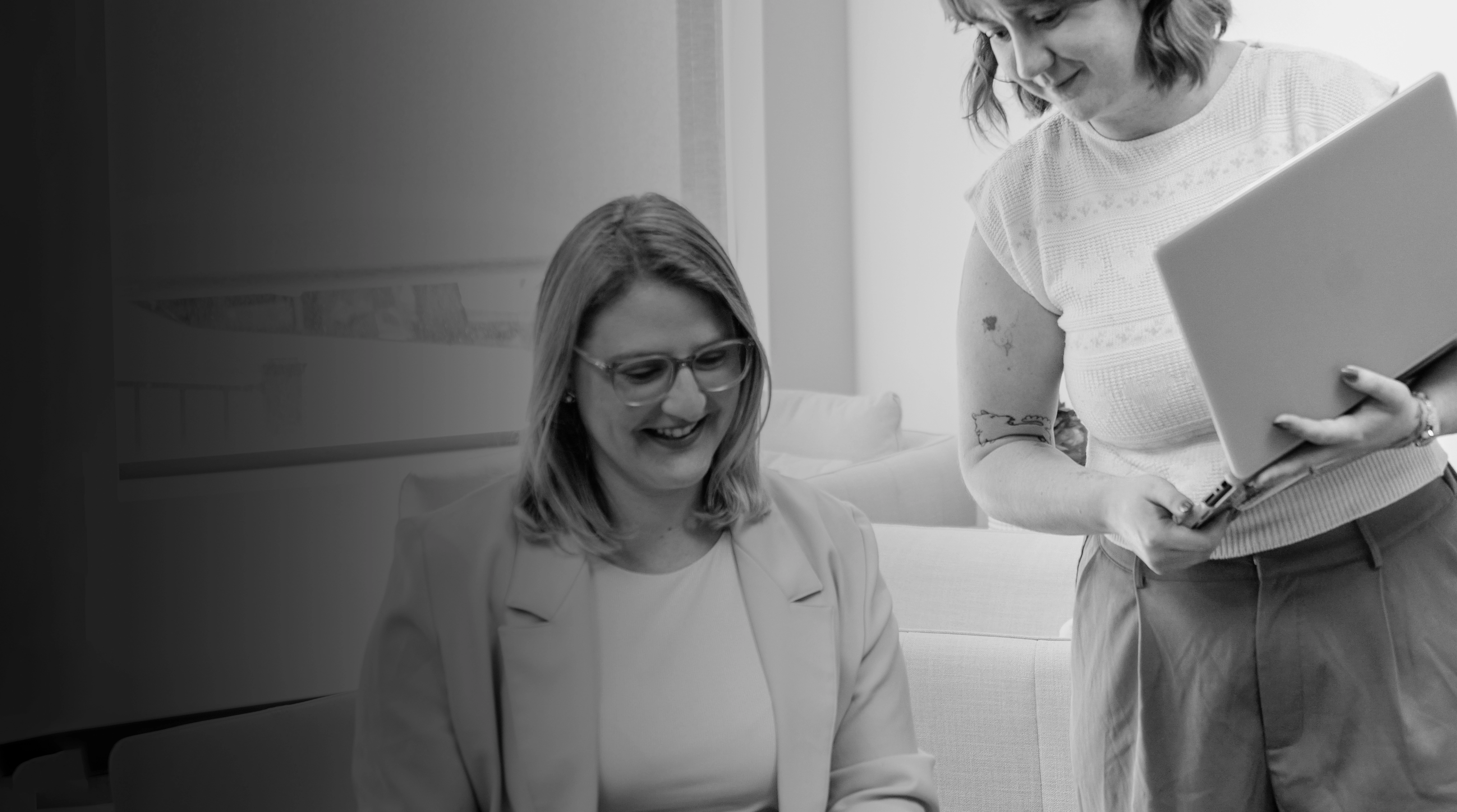 Two women collaborating in an office with one holding a laptop and the other smiling seated.
