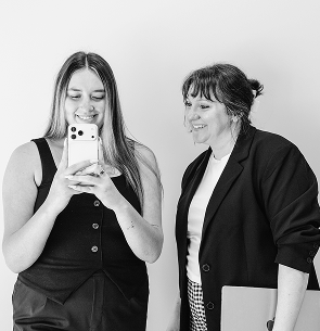 Two women smiling, one holding a smartphone taking a selfie, the other holding a laptop.