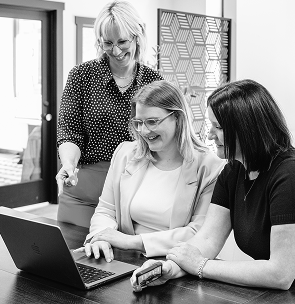 Three women smiling and working together on a laptop at a table in an office.
