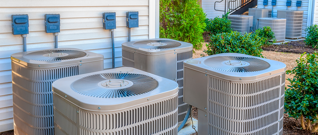 Four outdoor HVAC air conditioning units installed on a concrete pad next to a building, surrounded by green bushes and lawn.