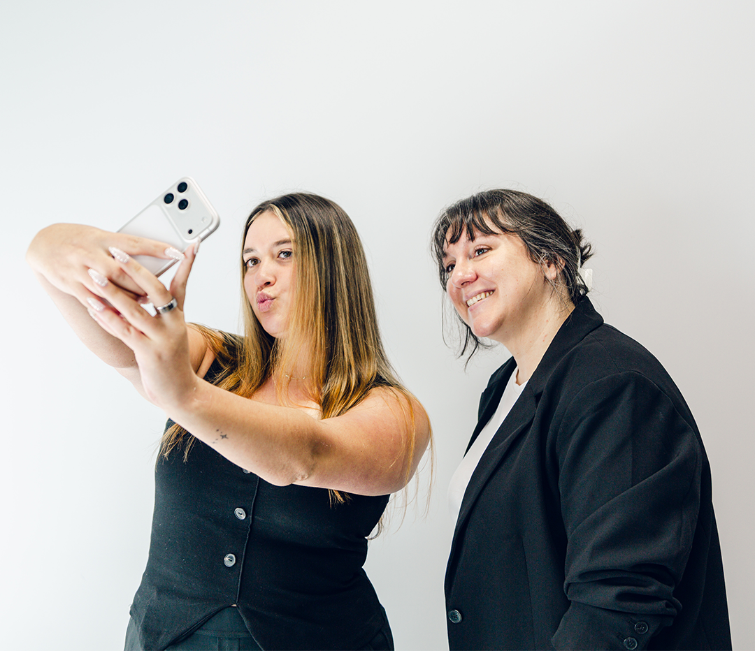 Two women taking a selfie together against a plain white background; the woman on the left is puckering her lips while holding a smartphone, and the woman on the right is smiling, wearing a black blazer over a white top.