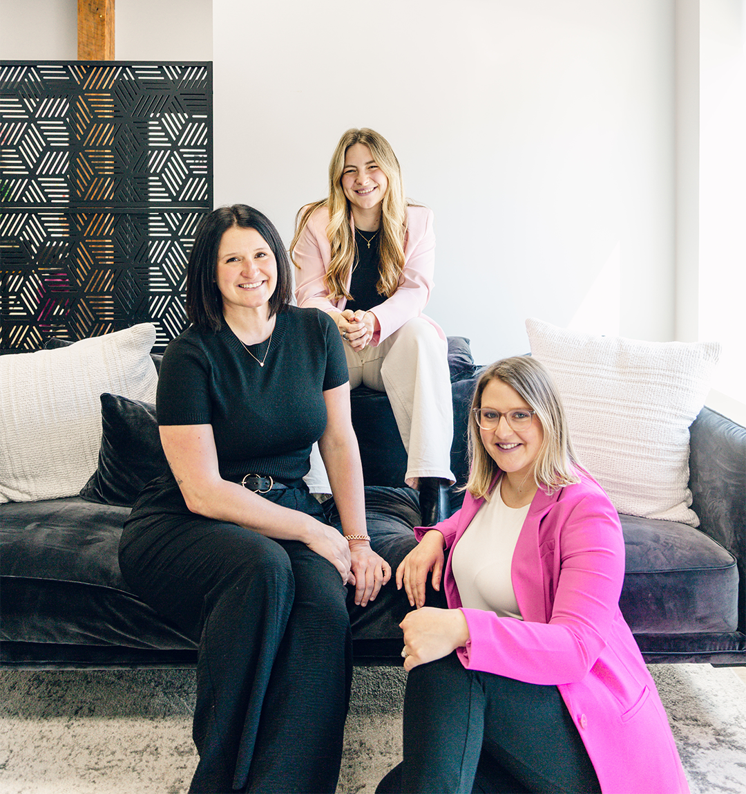 Three women smiling and posing in a bright, modern room; one in black dress sitting on a dark couch, one in a pink blazer and glasses sitting on the floor, and one with long blonde hair in a light pink blazer sitting on the couch back.