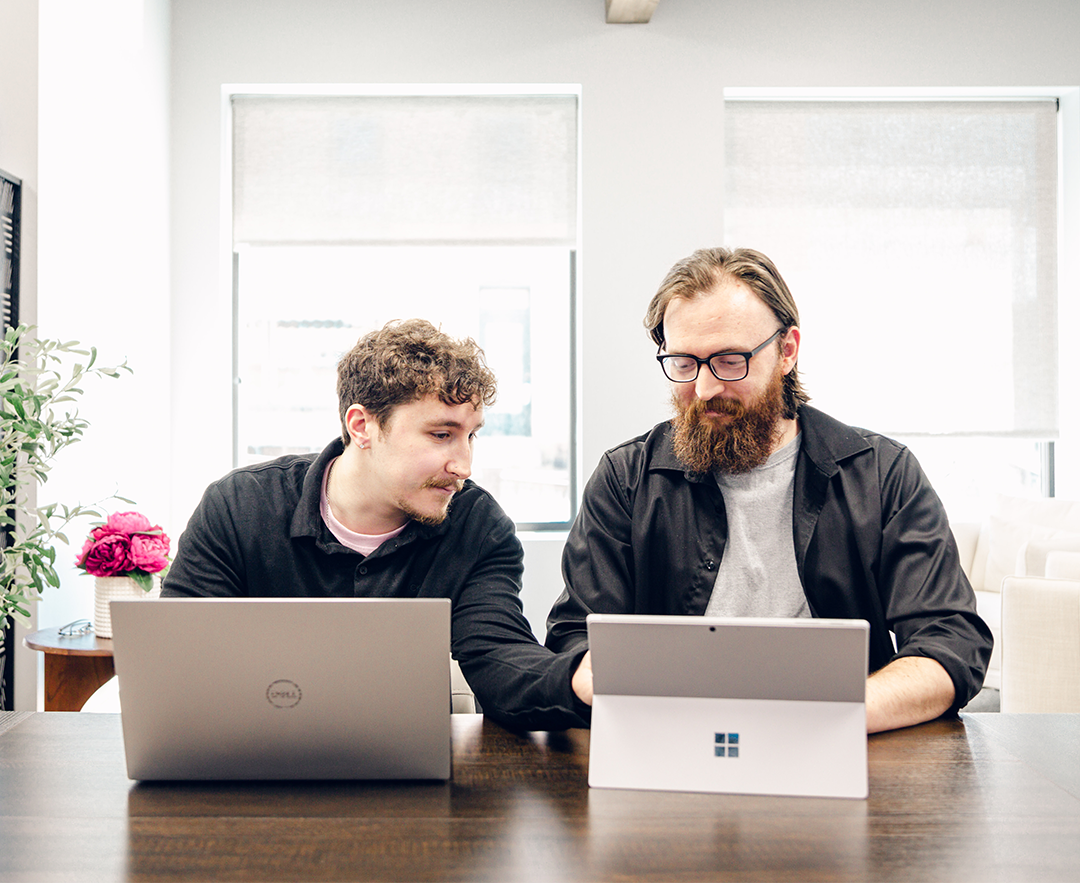 Two men sitting side by side at a wooden table in a bright room, working on laptops; the man on the left looks at the laptop of the man on the right who has a beard and glasses.