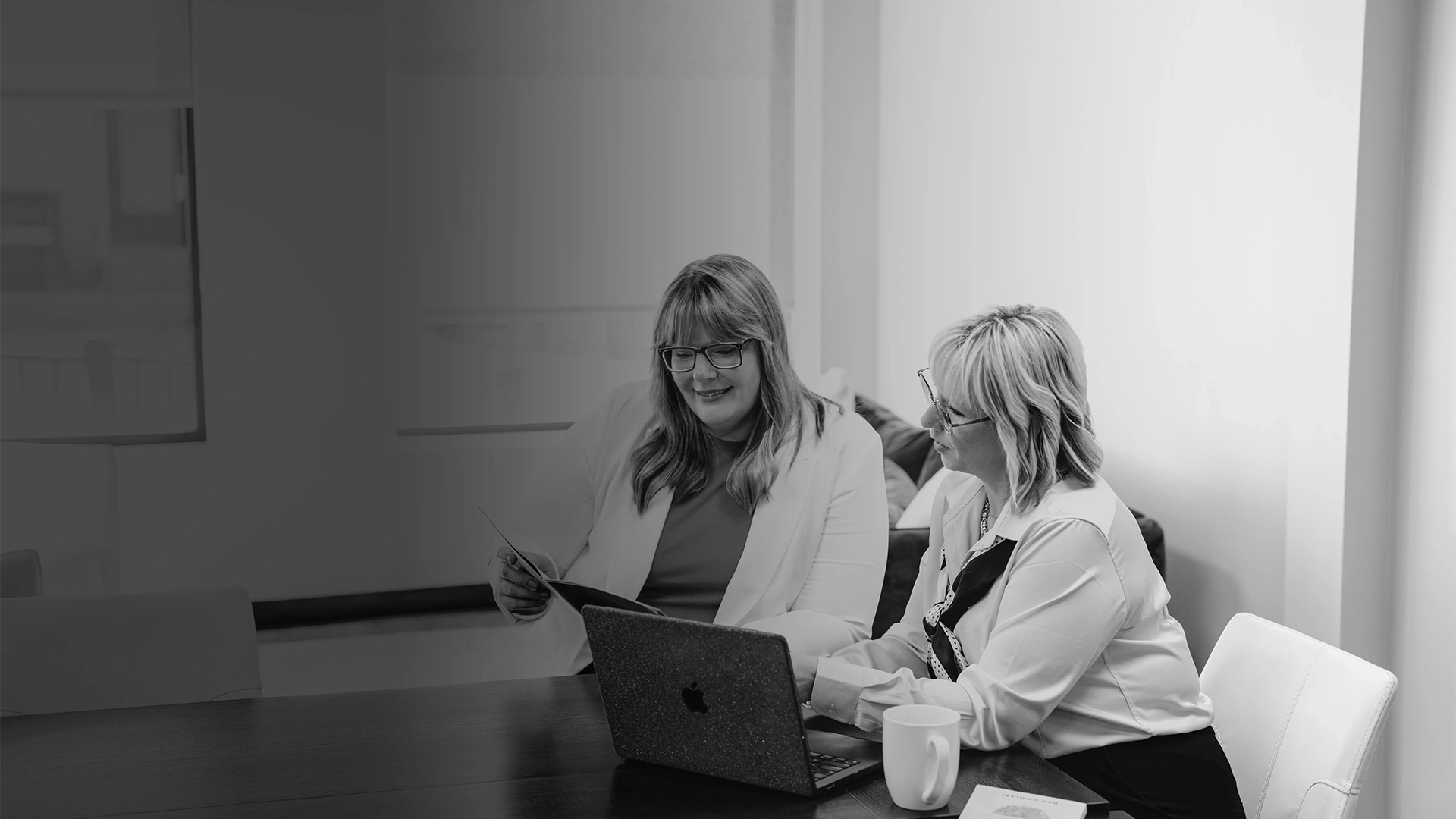 Two women collaborating in an office with one holding a laptop and the other smiling seated.