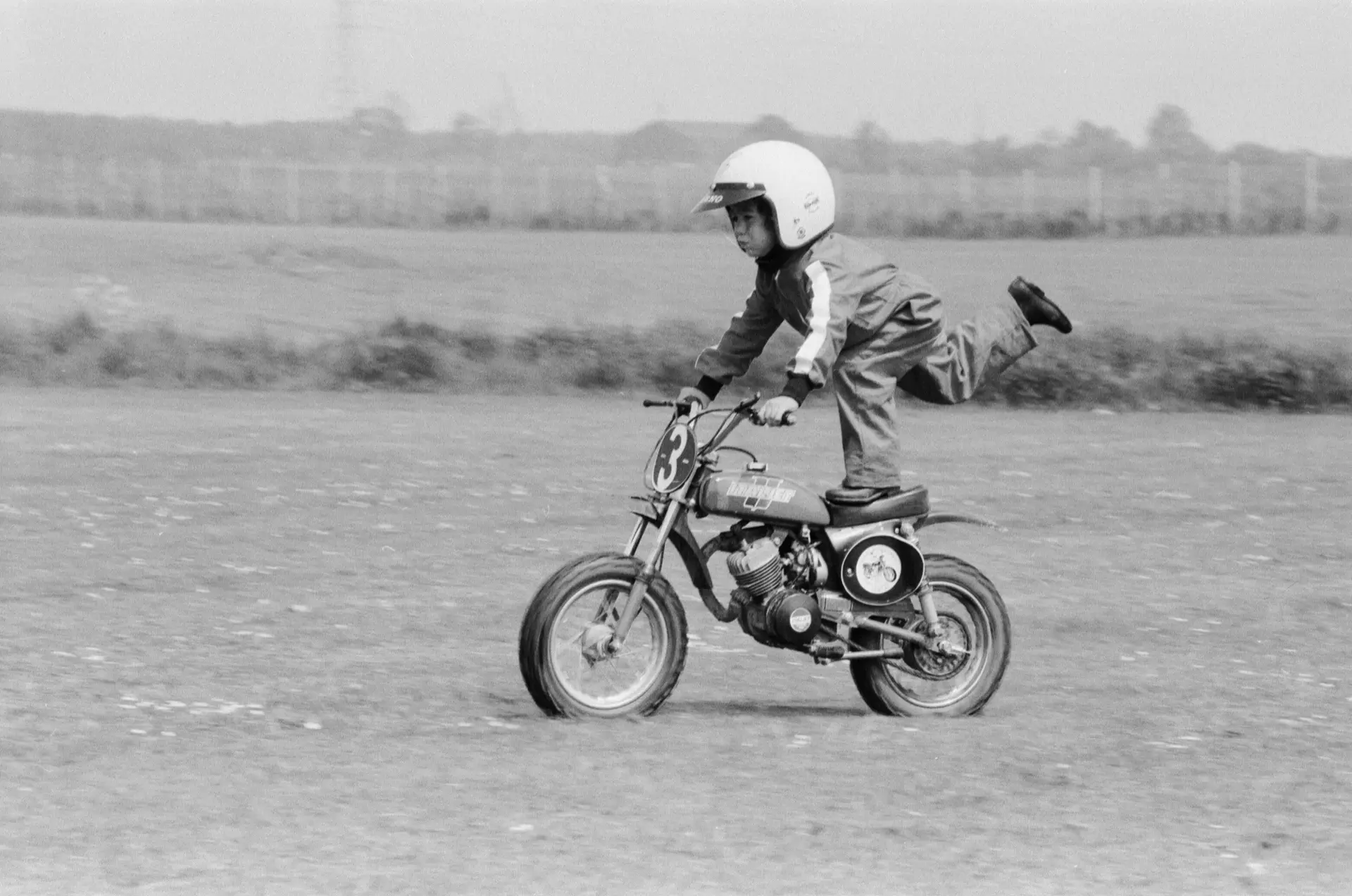 Black and white photo of a young child wearing a helmet riding a small motorbike with one leg extended backward on a grassy field.