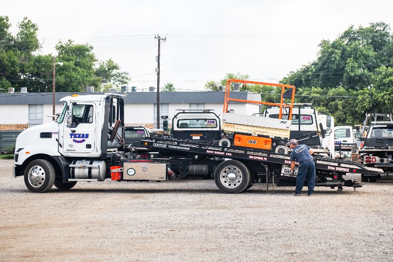 Tow truck operator securing a JLG scissor lift onto a flatbed tow truck in a parking lot with multiple service vehicles.