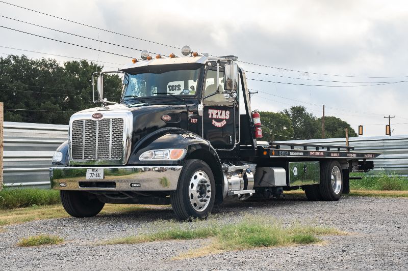 Black Texas Towing flatbed truck parked on gravel lot with chrome accents, metal fencing, green trees, and overcast sky in background.