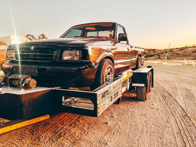 Black pickup truck loaded on flatbed trailer at sunset on dirt road, winch visible, towing equipment ready for transport.