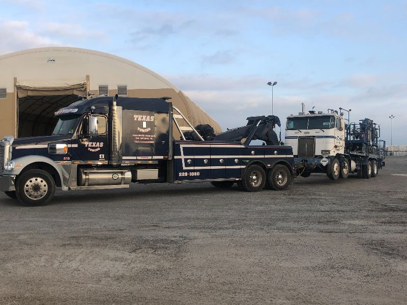 White flatbed tow truck parked on residential street, hauling a teal vintage classic car, sunny day with trees.