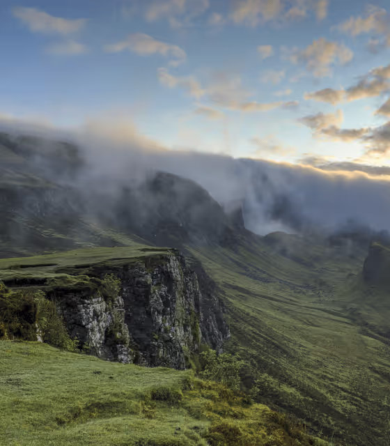 A green hillside with a rocky cliff and a cloudy sky.