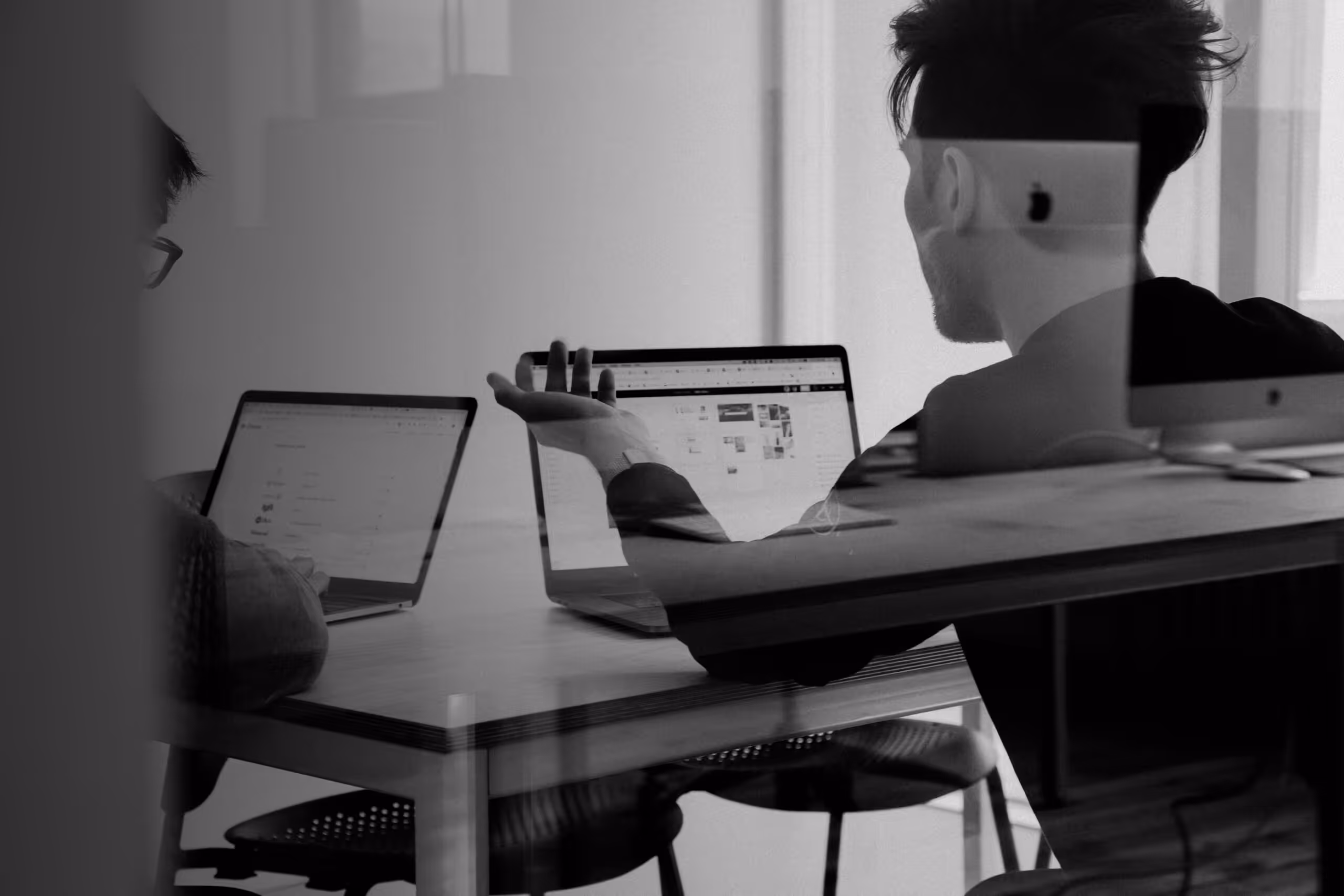 Two people working on laptops at a table, one gesturing while discussing, seen through a glass partition.