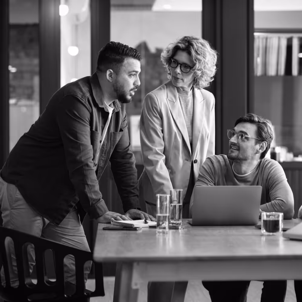 Three colleagues engaged in a discussion around a table with laptops and glasses of water in an office setting.