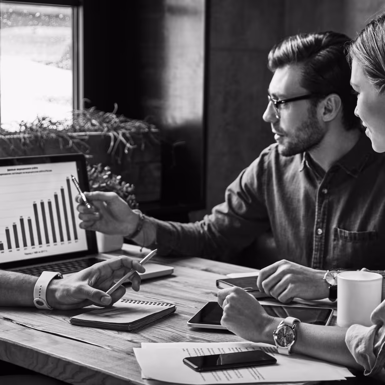 Three people collaborating at a wooden table discussing a laptop displaying a bar graph.