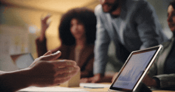 Hands of people in a business meeting gesturing towards a tablet displaying charts on a table.