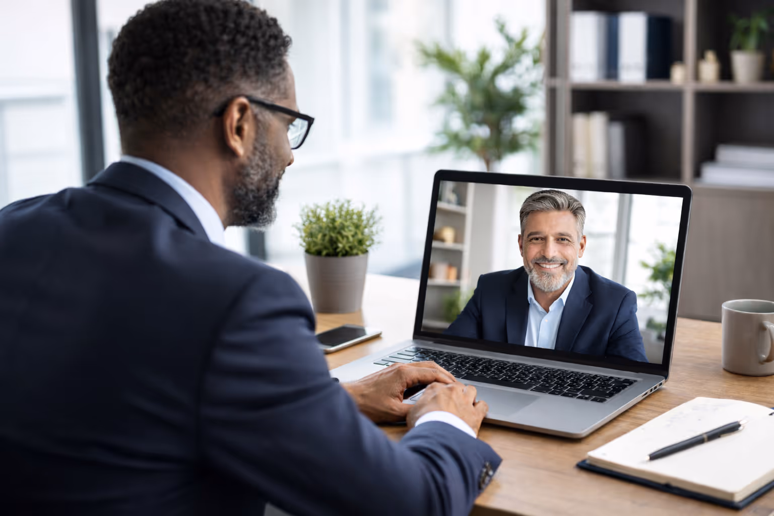 Two men in business suits having a video conference on a laptop in a modern office setting.