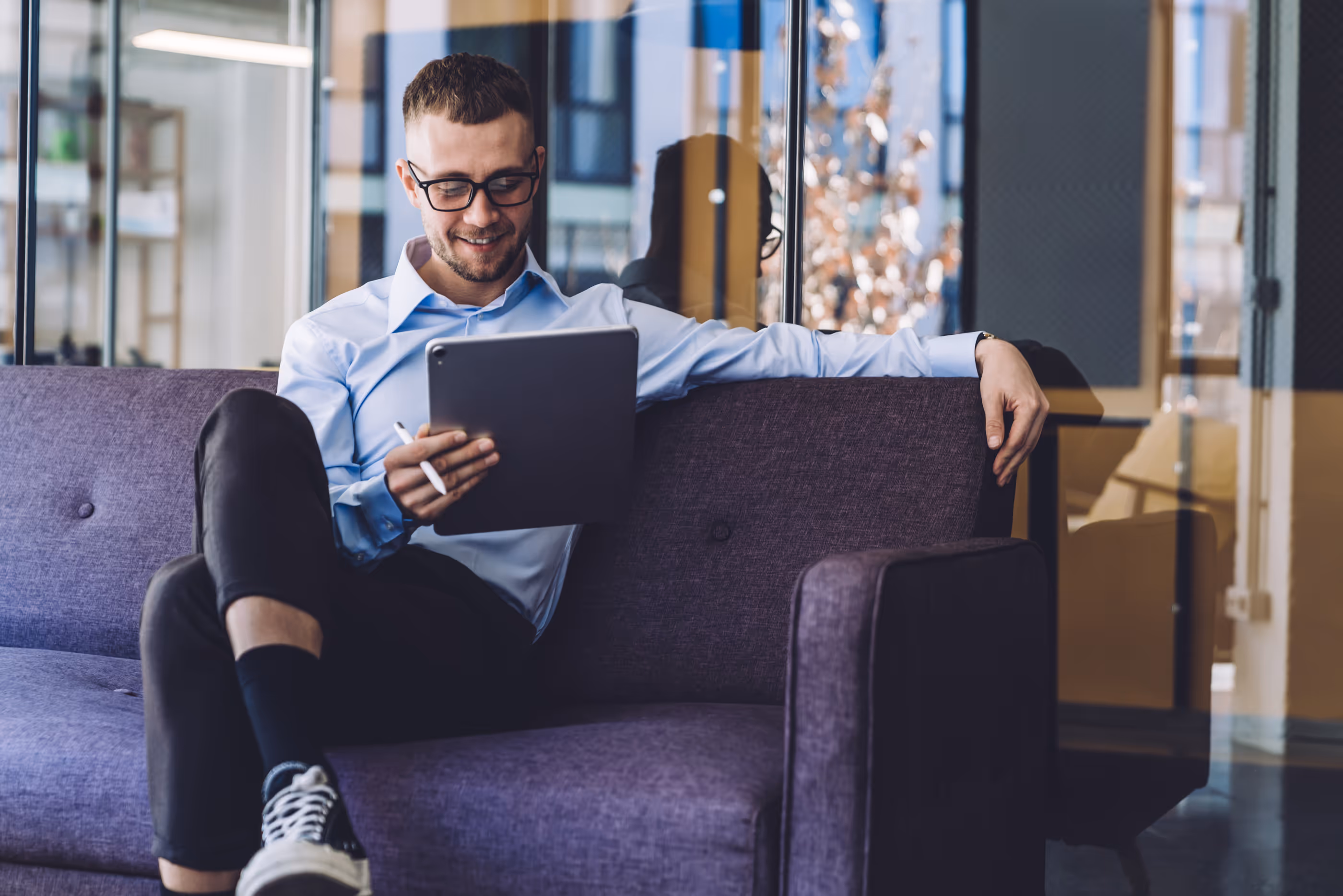 Smiling man with glasses sitting on a purple couch using a tablet with a stylus in a modern office.