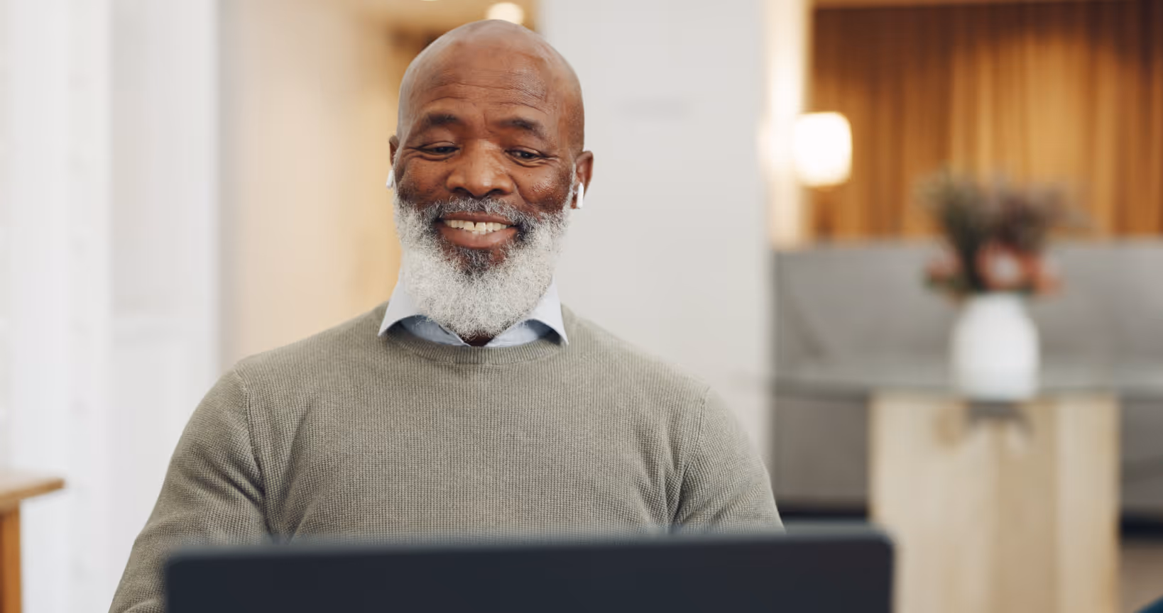 Smiling bearded man wearing earbuds looking at a laptop in a bright indoor setting.