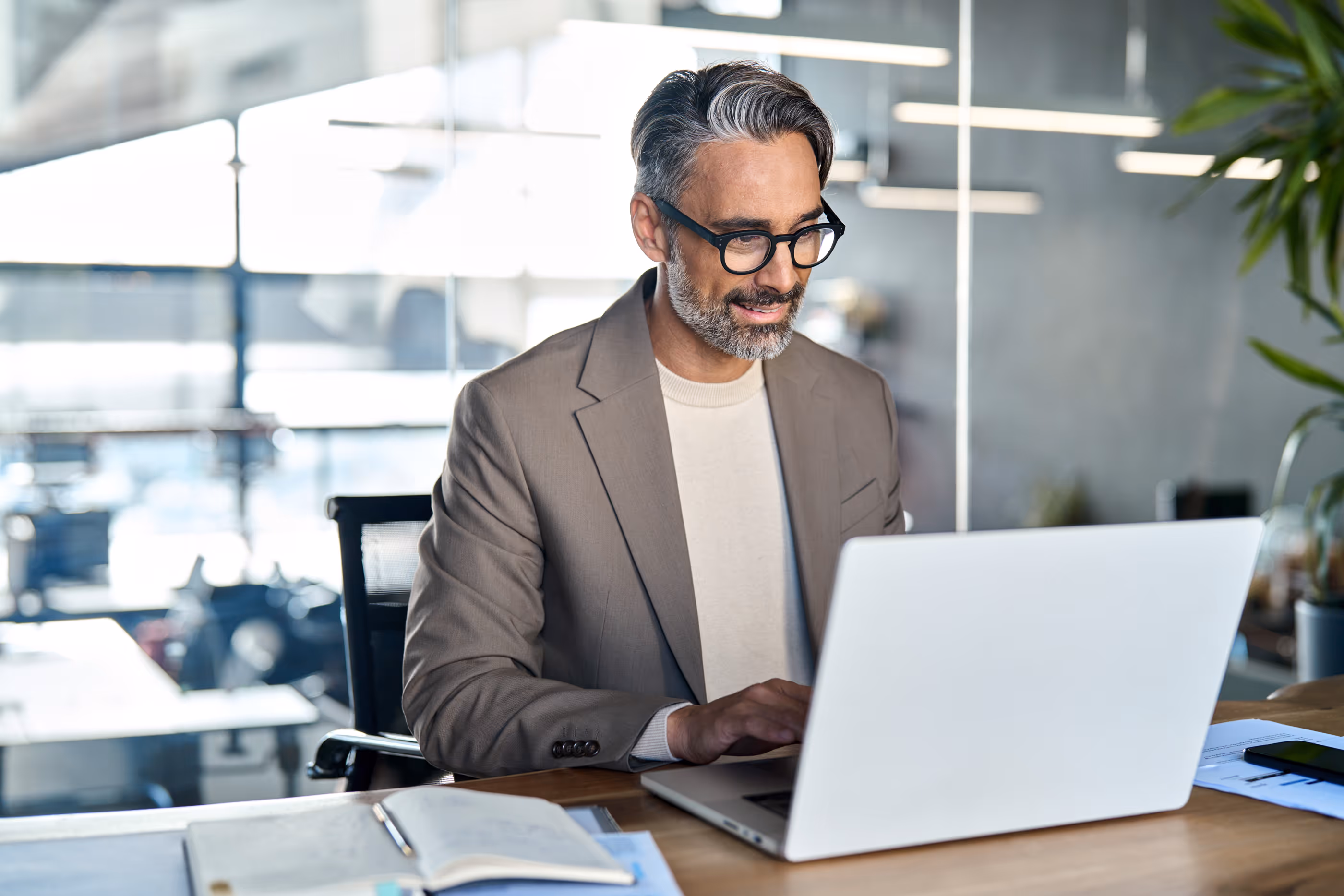 Man with gray hair and glasses, dressed in a brown blazer and beige sweater, working on a laptop in a modern office.
