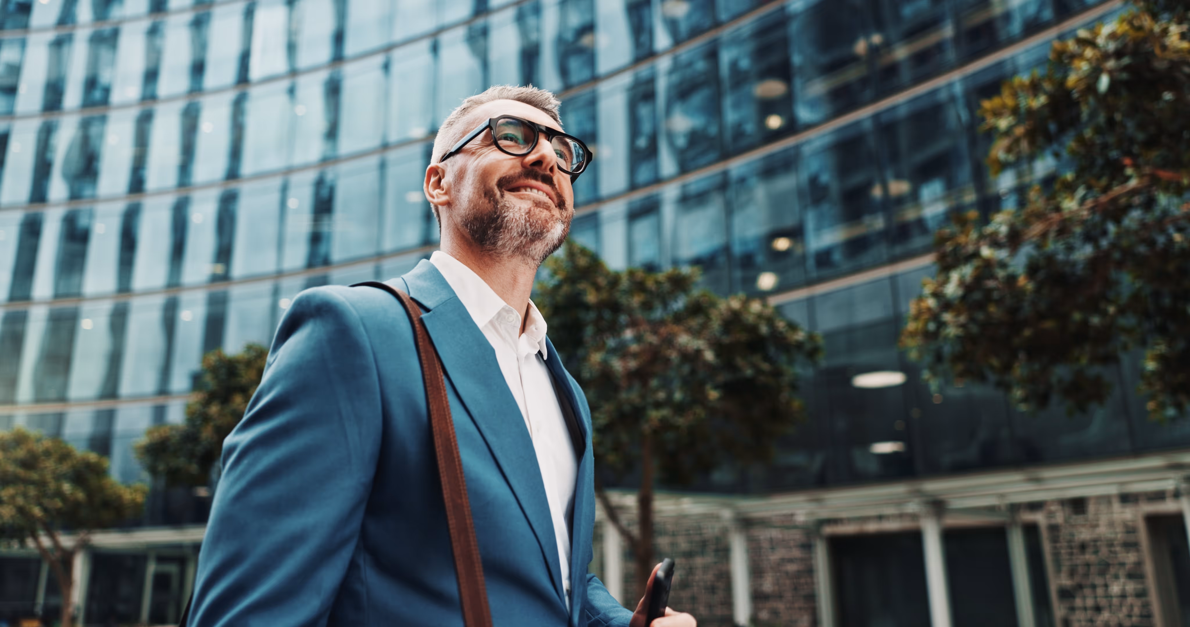 Smiling businessman in blue suit and glasses carrying a shoulder bag walking outside a modern glass office building.