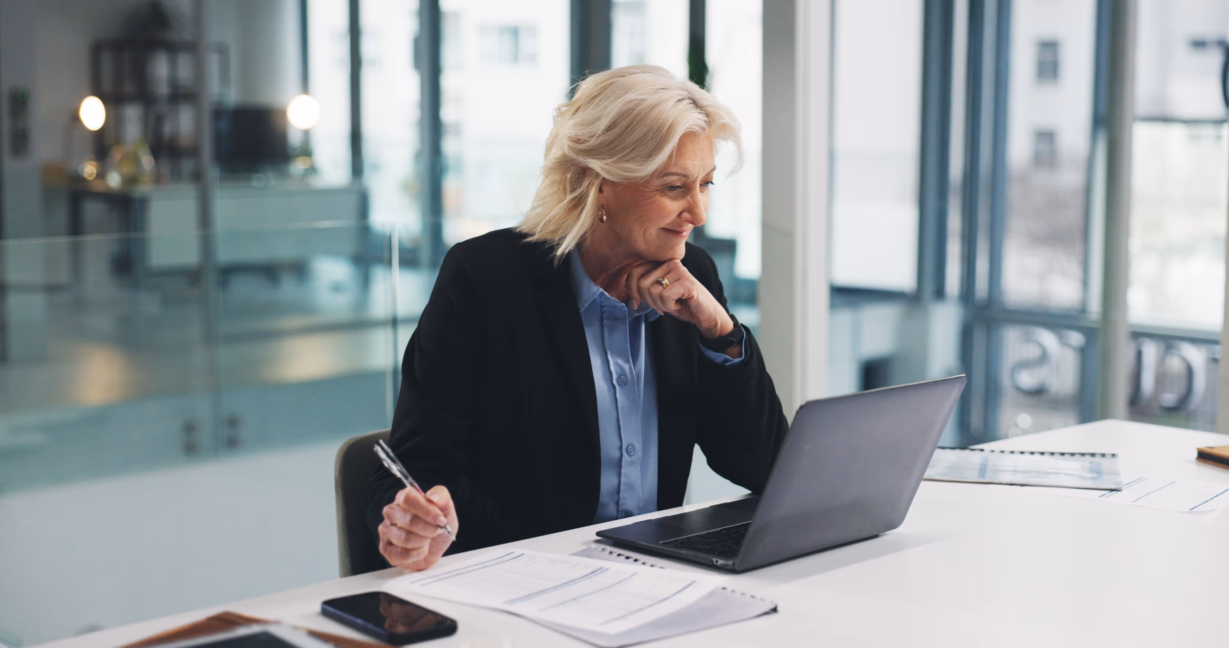 Middle-aged woman in business attire working on a laptop at a white desk with documents and a smartphone.