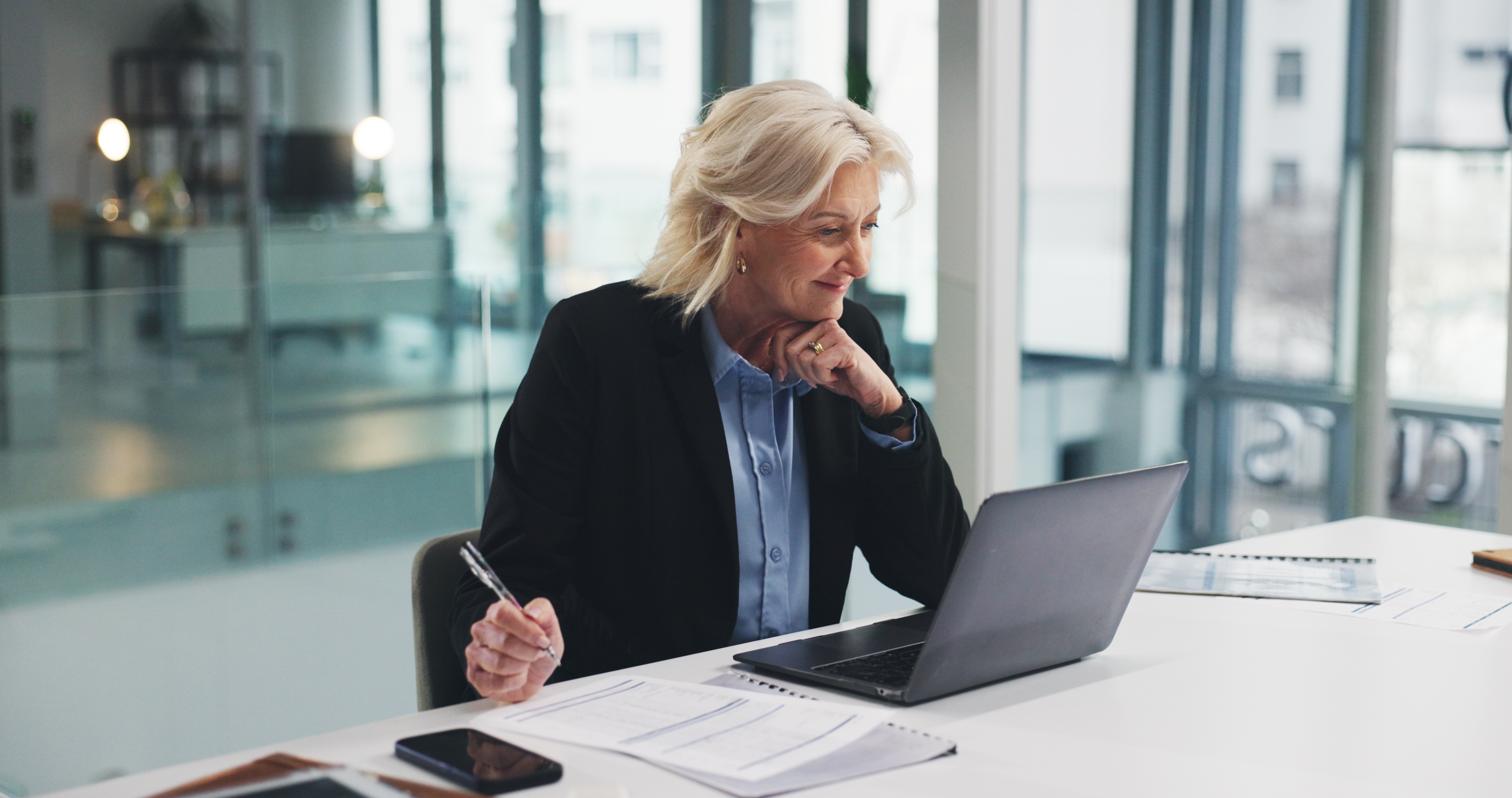 Middle-aged woman in business attire working on a laptop at a white desk with documents and a smartphone.