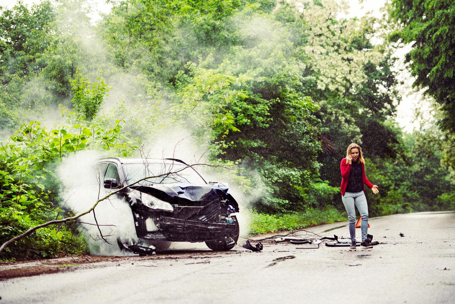 A woman stands next to a car that has been crushed by a tree, displaying a look of shock and concern.