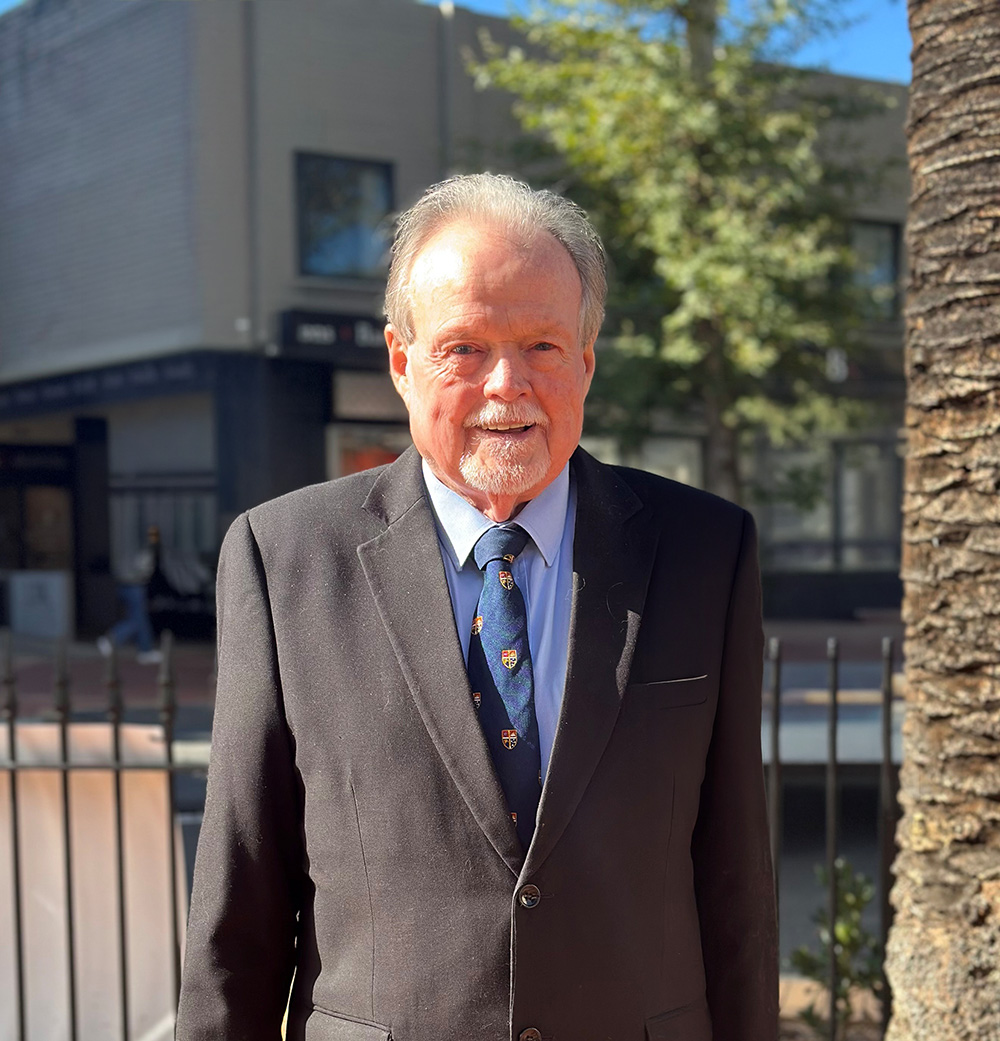 Older man with gray hair and goatee wearing a black suit and patterned tie outdoors in sunlight.