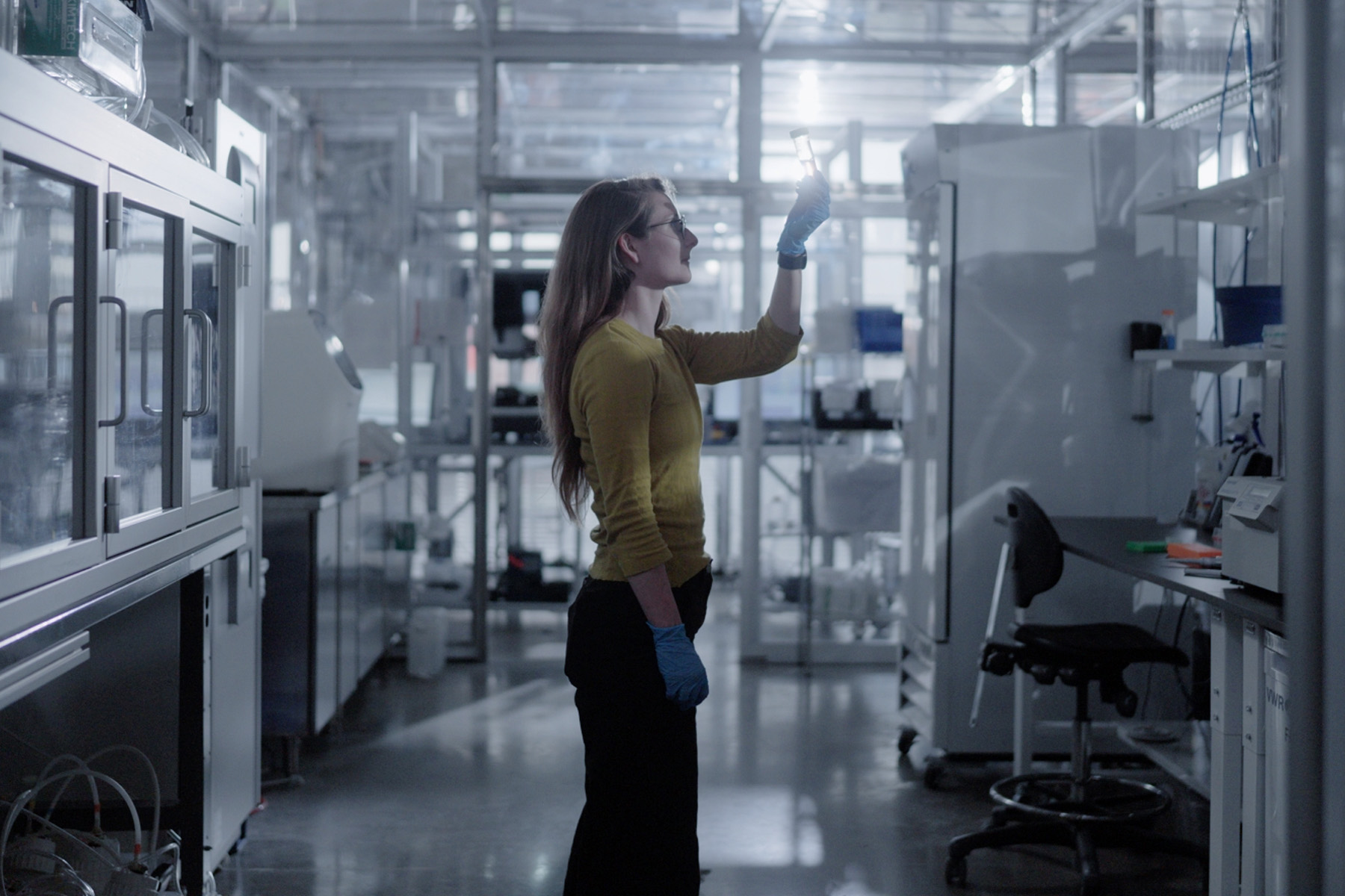 Scientist in a laboratory wearing blue gloves and holding up a vial to examine it closely.