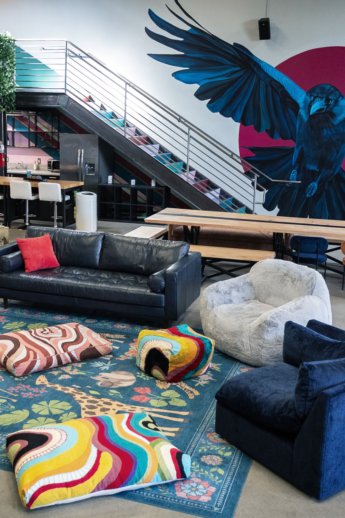 Modern lounge area with black leather sofa, blue and white armchairs, colorful cushions on a patterned rug, wooden tables, and a large blue bird mural on the wall under a staircase.