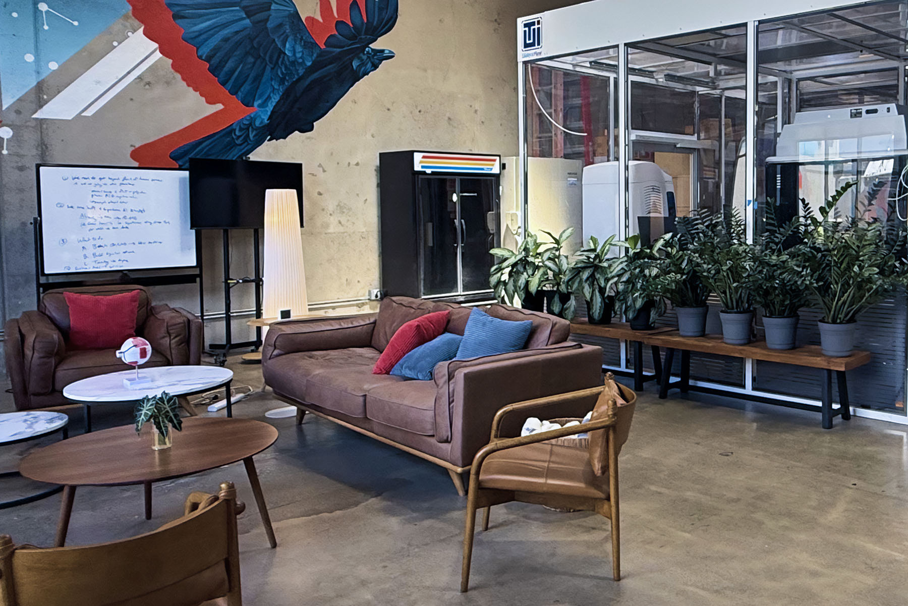 Modern office lounge with brown leather sofa and chair, colorful cushions, wooden tables, potted plants, a whiteboard, and a tall lamp under a wall mural of a bird.