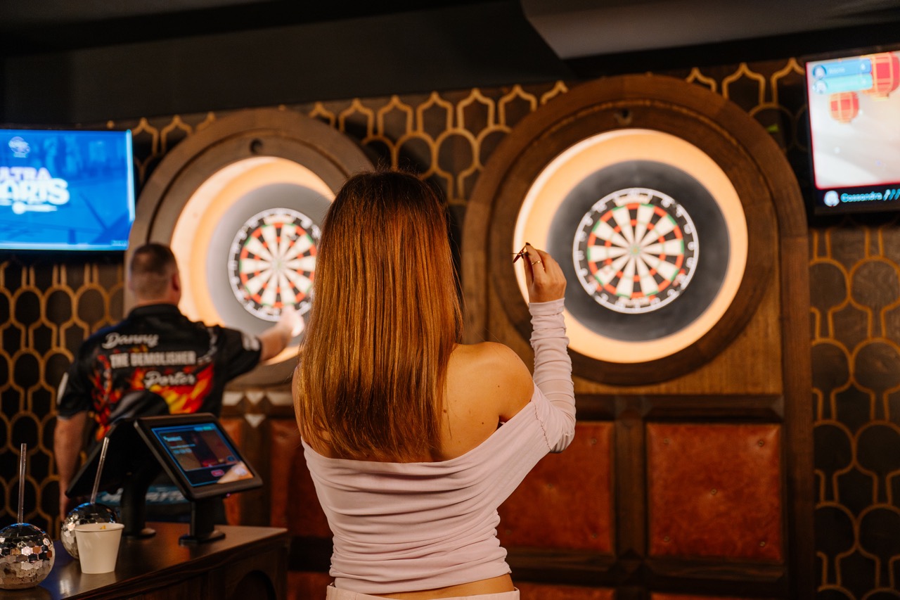 Two people playing darts indoors, aiming at illuminated dartboards mounted on a patterned wall.
