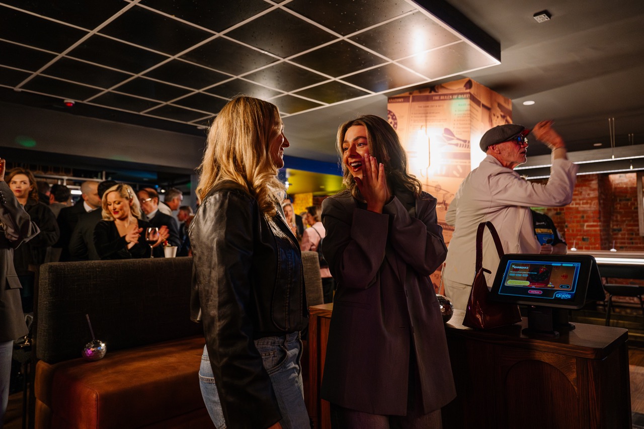 Two women laughing and talking in a dimly lit social gathering with other people in the background.