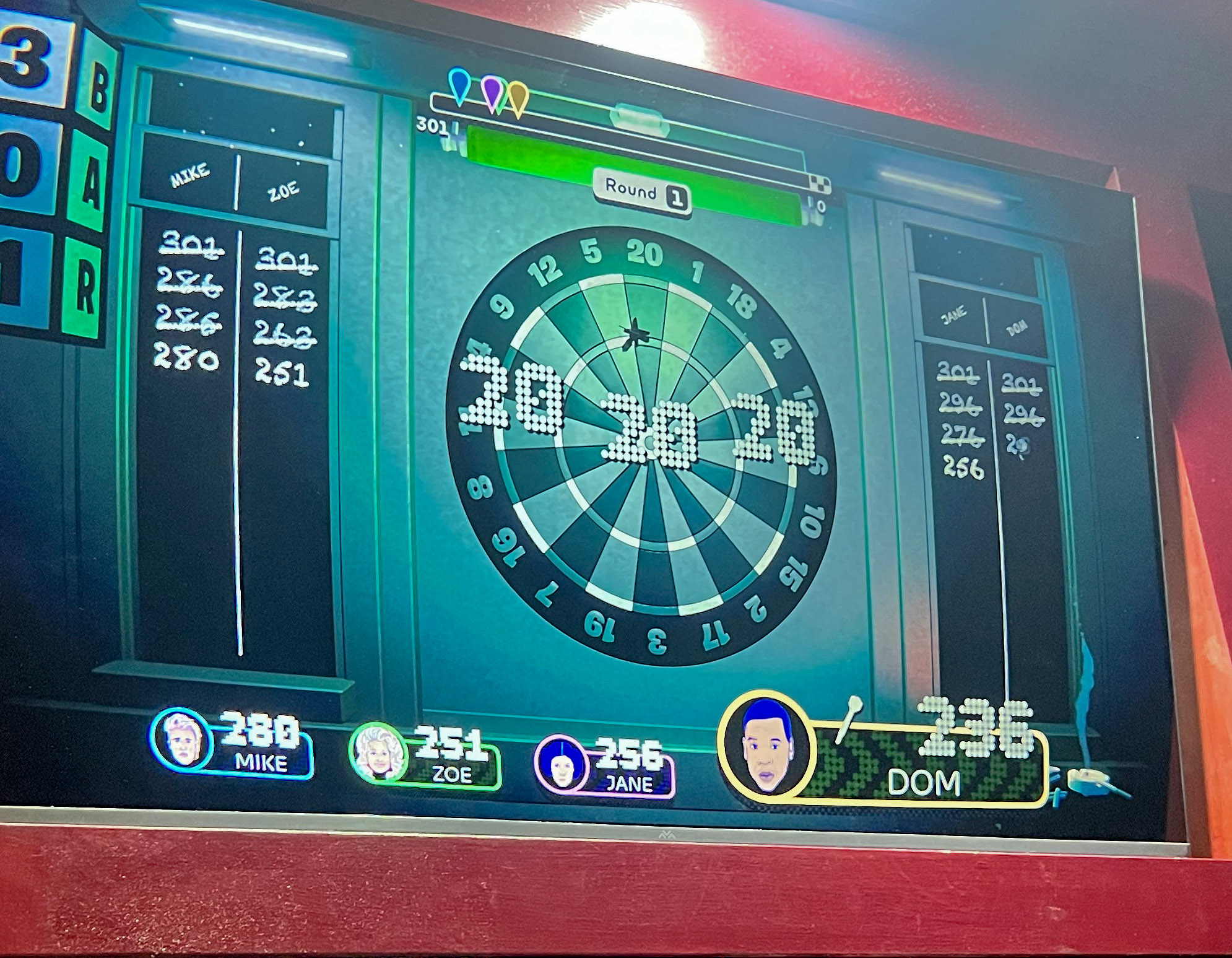 Electronic dartboard screen showing scores for players Mike, Zoe, Jane, and Dom in Round 1 of a 301 darts game.