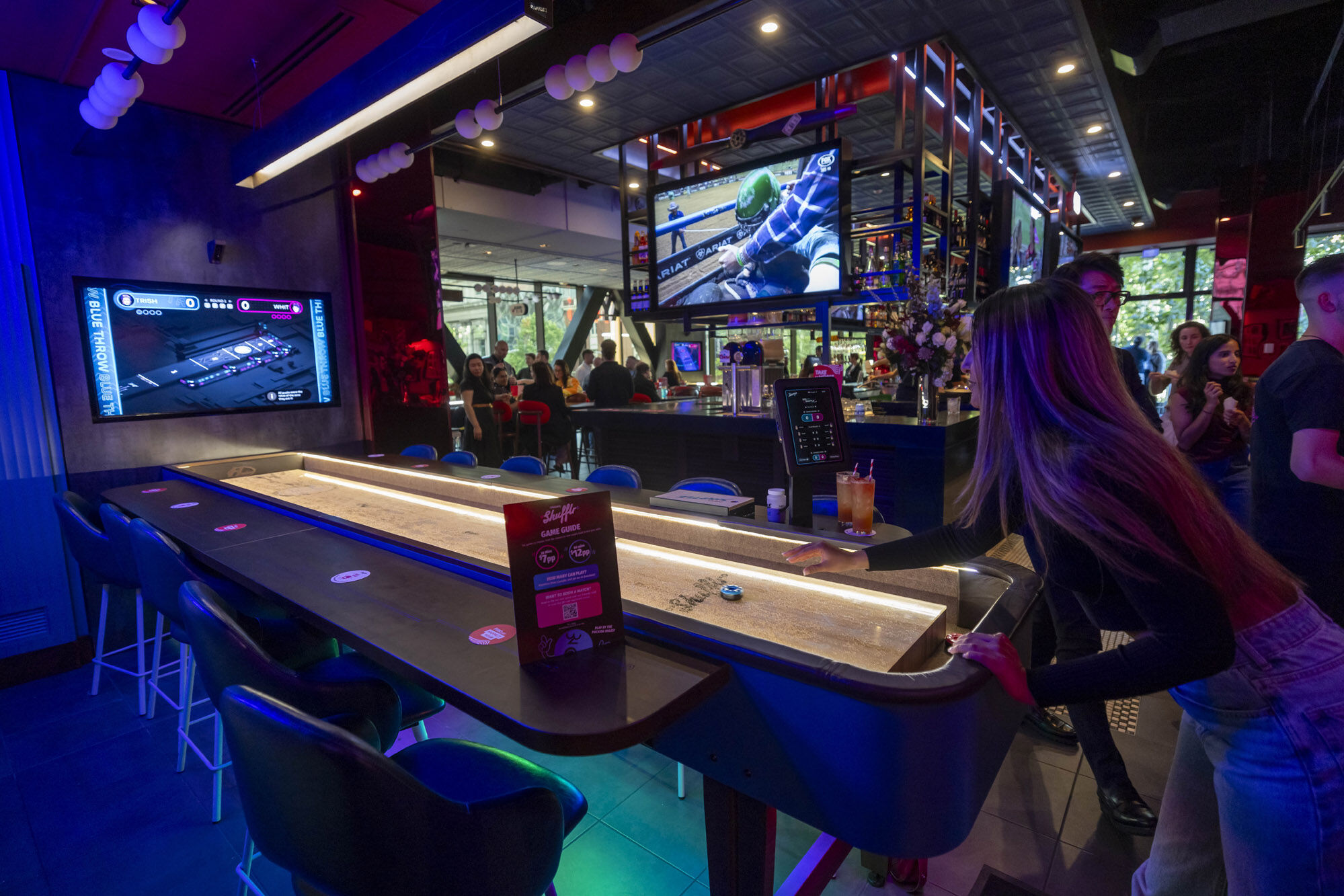 A woman playing Neoshuffle shuffleboard in a lively bar with multiple screens showing sports and games, surrounded by seated and standing patrons.
